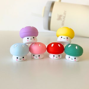 May include: Seven colourful mushroom-shaped objects with smiling faces. The mushrooms have caps in various colours, including purple, blue, pink, red, yellow, and green. The stems are white. A cream-coloured Stanley tumbler is in the background.