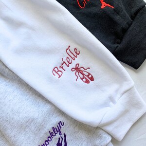 May include: Three sweatshirt sleeves with different colours and embroidery. The top sleeve is black with red embroidery that says "Chessa" and a red dancer silhouette. The middle sleeve is white with pink embroidery that says "Brielle" and a pair of pink ballet shoes. The bottom sleeve is grey with purple embroidery that says "Brooklyn" and a purple dancer silhouette.