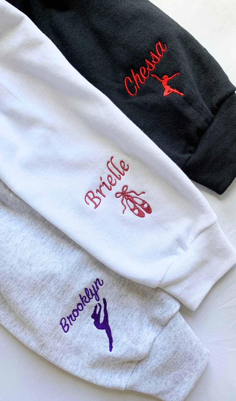 May include: Three sweatshirt sleeves with different colours and embroidery. The top sleeve is black with red embroidery that says "Chessa" and a red dancer silhouette. The middle sleeve is white with pink embroidery that says "Brielle" and a pair of pink ballet shoes. The bottom sleeve is grey with purple embroidery that says "Brooklyn" and a purple dancer silhouette.