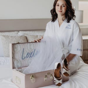 May include: A white waffle-knit robe with blue embroidery reading Mrs. Henche and 08.08.25, paired with a light pink rectangular storage case featuring the name Lexie in blue script, containing a white veil and a pair of white strappy heels.