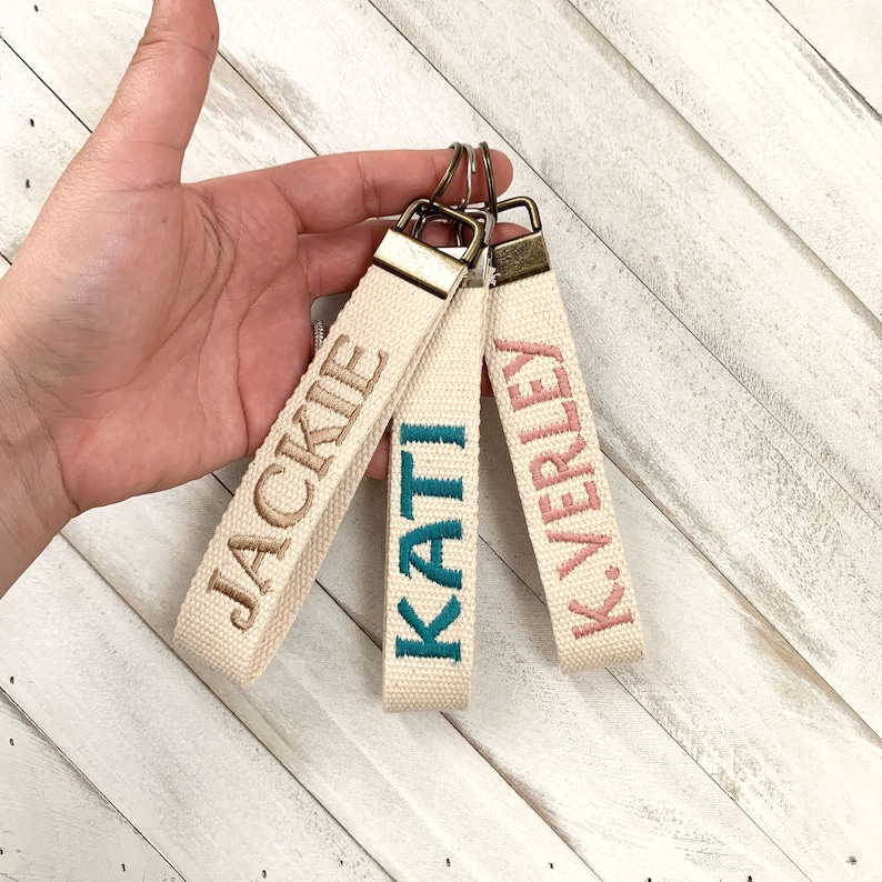 May include: Three personalized canvas keychains with embroidered names: "JACKIE" in brown, "KATI" in teal, and "K. VERLEY" in pink. Each keychain has a beige strap, a metal clasp, and a metal ring for keys. The keychains are displayed against a white wooden background.