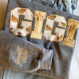 May include: Gray sweatshirt with a large embroidered patch that says "GIGI" with a Winnie the Pooh design. The patch has a white background with yellow butterflies. The sweatshirt also has a smaller embroidered patch on the sleeve that says "Bennett Blake".