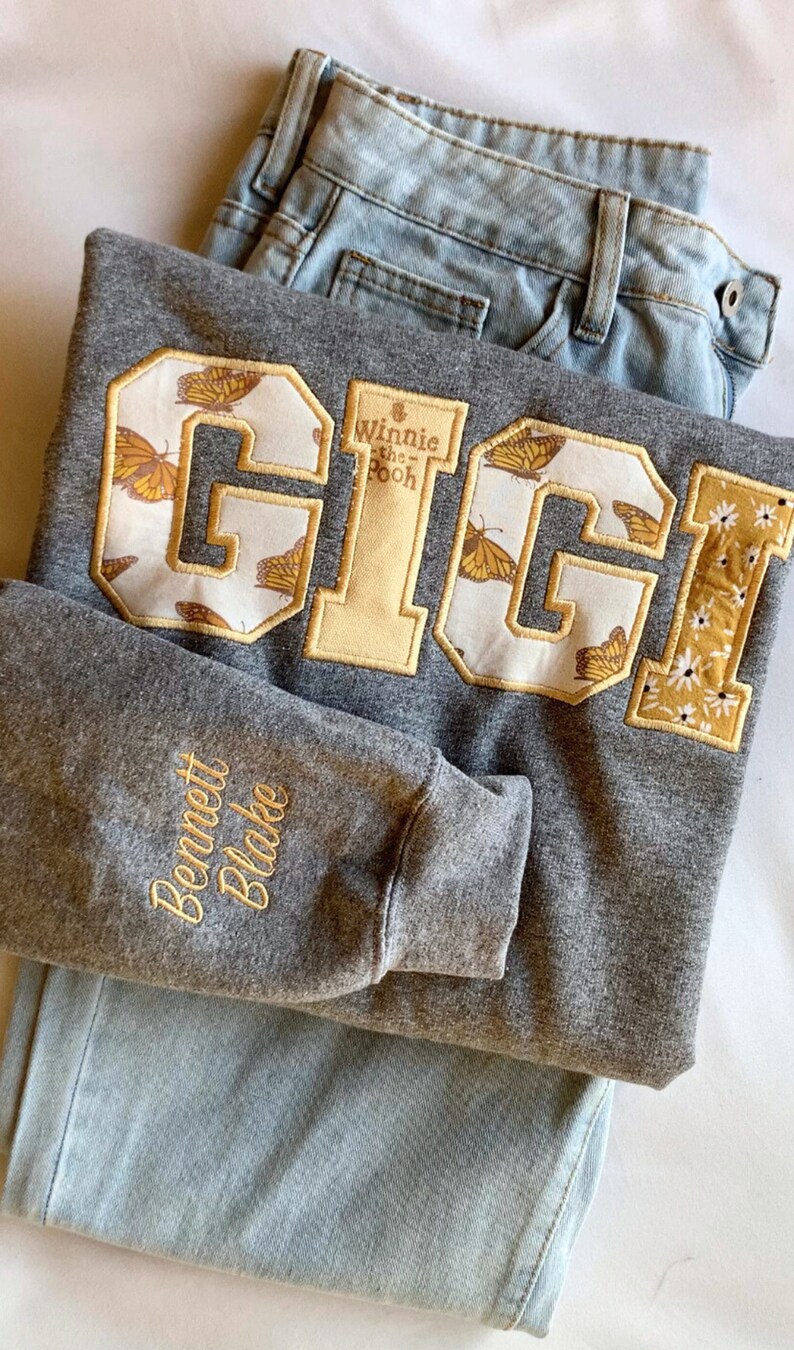 May include: Gray sweatshirt with a large embroidered patch that says "GIGI" with a Winnie the Pooh design. The patch has a white background with yellow butterflies. The sweatshirt also has a smaller embroidered patch on the sleeve that says "Bennett Blake".