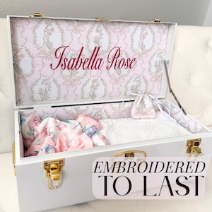 Pode incluir: Um baú de recordações branco com fechos e dobradiças dourados. A tampa interior apresenta um padrão de fitas e flores cor-de-rosa e brancas com o nome Isabella Rose bordado em rosa. Dentro há uma pequena bolsa e tecido floral.