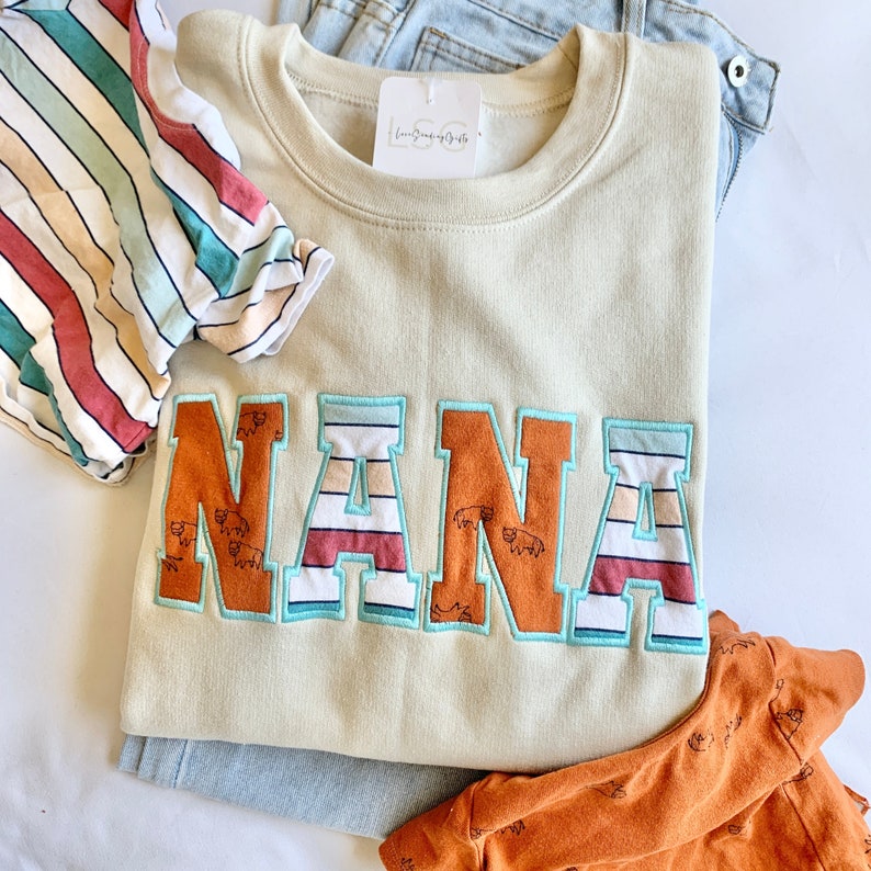 May include: A light beige sweatshirt with the word "NANA" in large, colorful, embroidered letters. The letters are outlined in blue and have stripes of red, white, and blue.