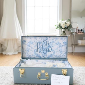 May include: A light blue storage trunk with gold-toned metal latches and a handle. The interior lid features a blue and white floral toile pattern with the embroidered monogram ACS. A white card with handwritten text rests in front of the trunk.