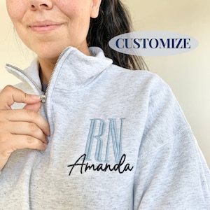 May include: Grey zipped sweatshirt with a blue embroidered "RN" monogram and the name "Amanda" in black script.