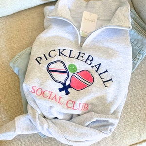 May include: Gray sweatshirt with a white embroidered design of two pickleball paddles and a ball. The text "Pickleball Social Club" is embroidered in blue and pink.