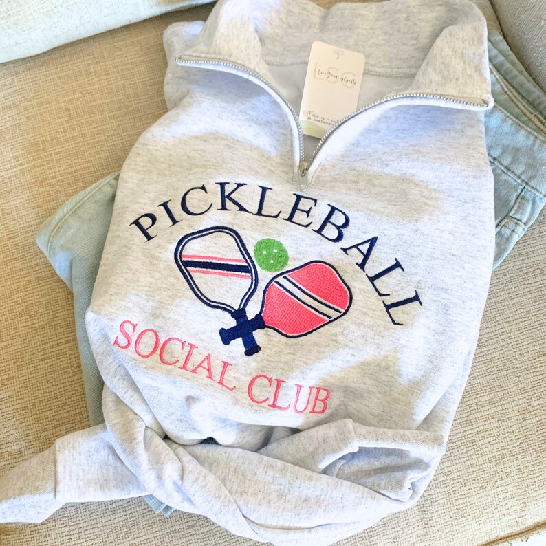 May include: Gray sweatshirt with a white embroidered design of two pickleball paddles and a ball. The text "Pickleball Social Club" is embroidered in blue and pink.