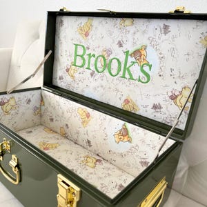 May include: An olive green metal storage trunk with gold-toned latches and handles. The interior is lined with a cream-colored fabric featuring Winnie the Pooh illustrations and the embroidered green name Brooks on the inside of the lid.