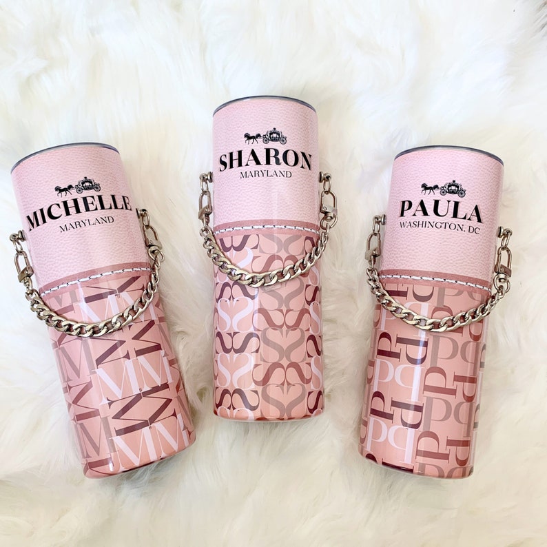 May include: Three pink personalised tumblers with a silver chain handle. The tumblers have a pink and brown patterned design and feature the names "Michelle", "Sharon", and "Paula" with their respective states.