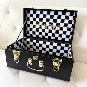 May include: A black rectangular storage chest with gold-toned metal latches and a central handle. The interior is lined with a black and white checkered pattern, and the name Brooks is printed in gold lettering on the inside of the lid.
