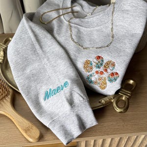 May include: A light gray crewneck sweatshirt featuring an embroidered floral paw print design on the chest in shades of orange, yellow, and teal. The sleeve is embroidered with the name Maeve in teal script lettering.