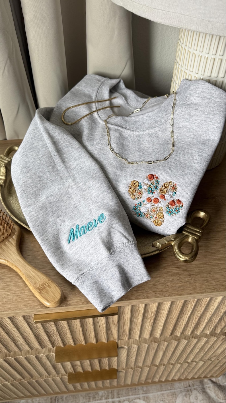 May include: A light gray crewneck sweatshirt featuring an embroidered floral paw print design on the chest in shades of orange, yellow, and teal. The sleeve is embroidered with the name Maeve in teal script lettering.