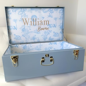 May include: A light blue storage trunk with gold-toned metal latches and a handle. The interior is lined with white fabric featuring a blue toile pattern of animals and trees. The name William Bowen is embroidered in gold thread on the lid interior.