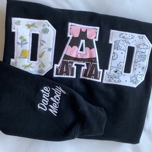 May include: Black sweatshirt with the word "DAD" in large white letters. Each letter is made of different patterned fabric. The sweatshirt has "Dante Melody" embroidered on the sleeve.