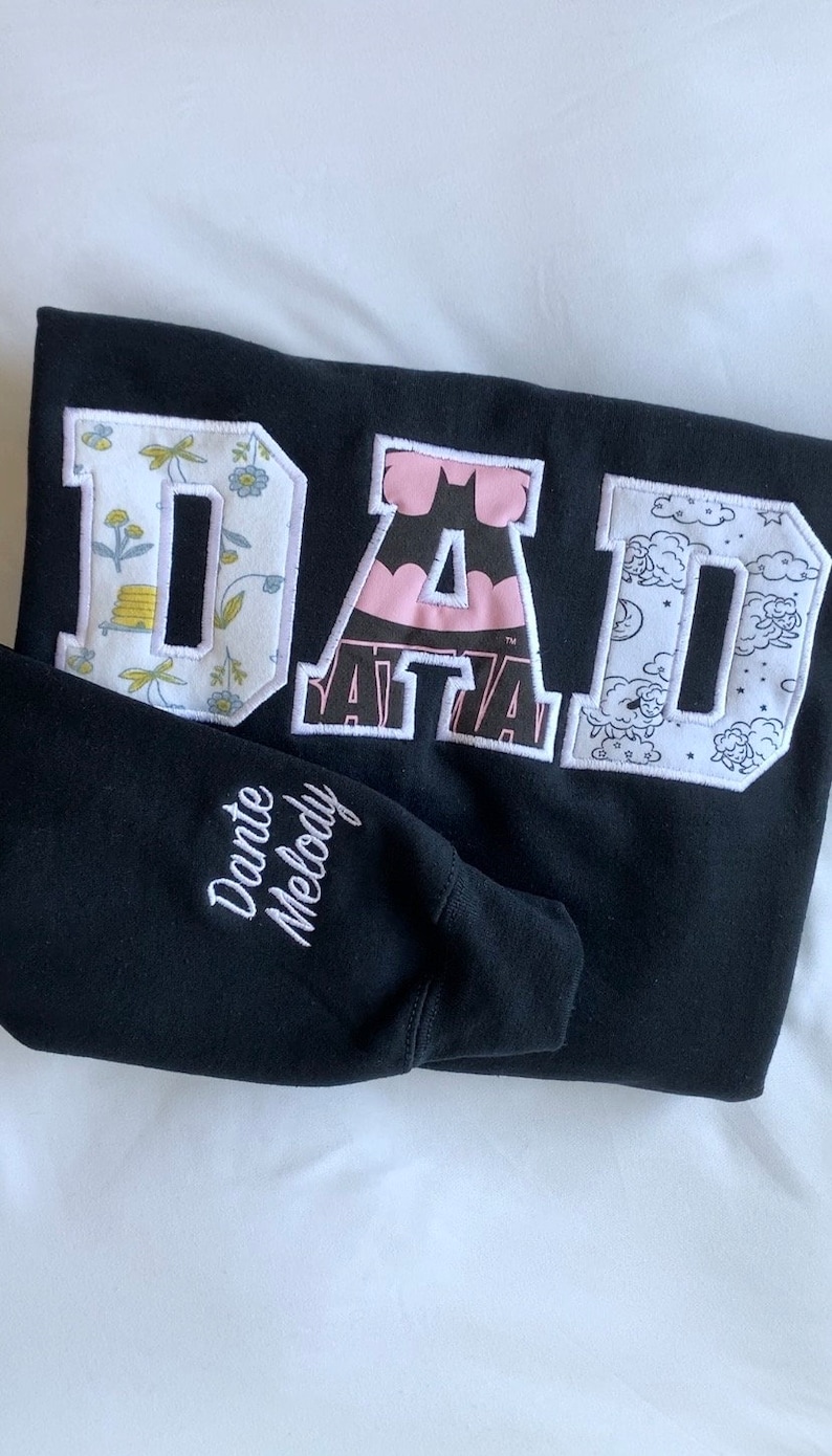 May include: Black sweatshirt with the word "DAD" in large white letters. Each letter is made of different patterned fabric. The sweatshirt has "Dante Melody" embroidered on the sleeve.