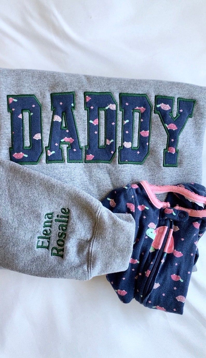 May include: Gray sweatshirt with the word "DADDY" embroidered in navy blue with a green outline. The sweatshirt has a small turtle pattern. The sleeve has the names "Elena Rosalie" embroidered in green.