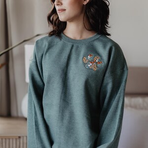 May include: A dark teal crewneck sweatshirt featuring an embroidered floral paw print design on the upper left chest. The right sleeve cuff displays the white embroidered text Bodhi Willow Ranger in a script font.