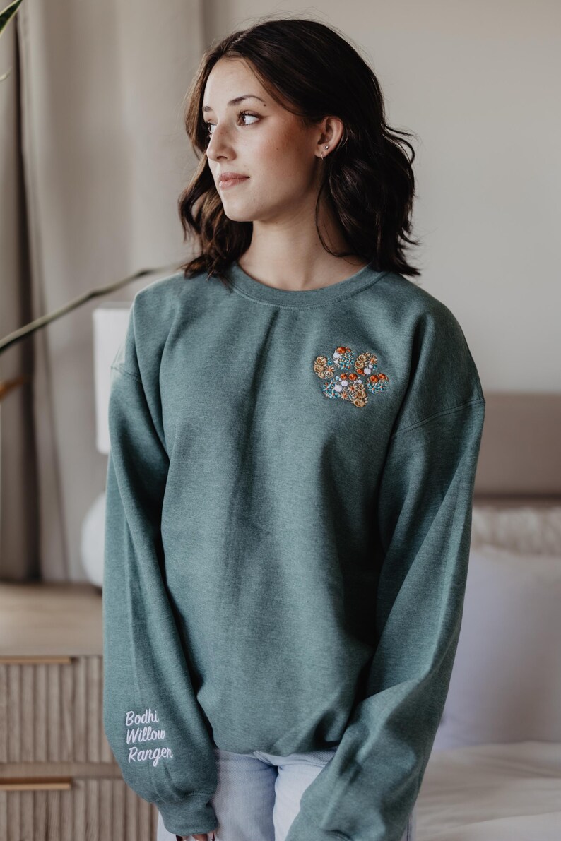 May include: A dark teal crewneck sweatshirt featuring an embroidered floral paw print design on the upper left chest. The right sleeve cuff displays the white embroidered text Bodhi Willow Ranger in a script font.