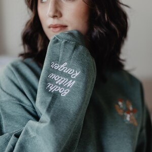 May include: A heathered sage green long-sleeve sweatshirt featuring white cursive embroidery on the sleeve that reads Bodhi Willow Ranger and a small, colorful embroidered graphic on the chest area depicting a cluster of mushrooms.
