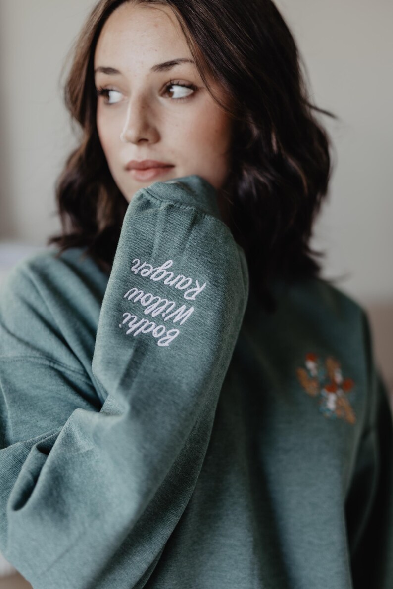 May include: A heathered sage green long-sleeve sweatshirt featuring white cursive embroidery on the sleeve that reads Bodhi Willow Ranger and a small, colorful embroidered graphic on the chest area depicting a cluster of mushrooms.