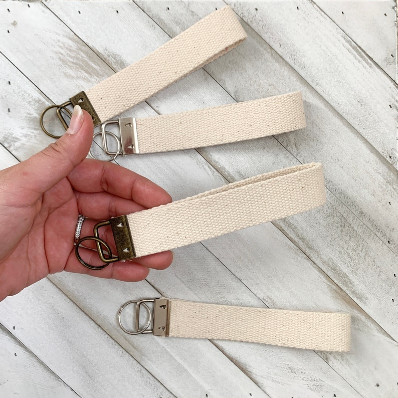 May include: Four cream-colored canvas key fobs with antique brass hardware and silver-tone key rings. The key fobs are made of a woven fabric and are arranged on a white wooden surface.