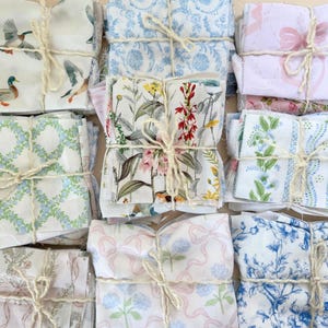 Puede incluir: Paquetes de cuadrados de tela atados con hilo, con varios patrones florales y de naturaleza. Los dise&ntilde;os incluyen patos, flores, lazos e impresiones florales azules. Las telas son en tonos de blanco, azul, rosa y verde.