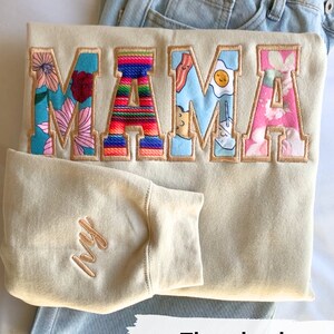 May include: A beige sweatshirt with the word "MAMA" embroidered in colorful patterned letters. The letters are outlined in golden brown thread. The sweatshirt has a small "Ivy" embroidered on the sleeve in golden brown thread.