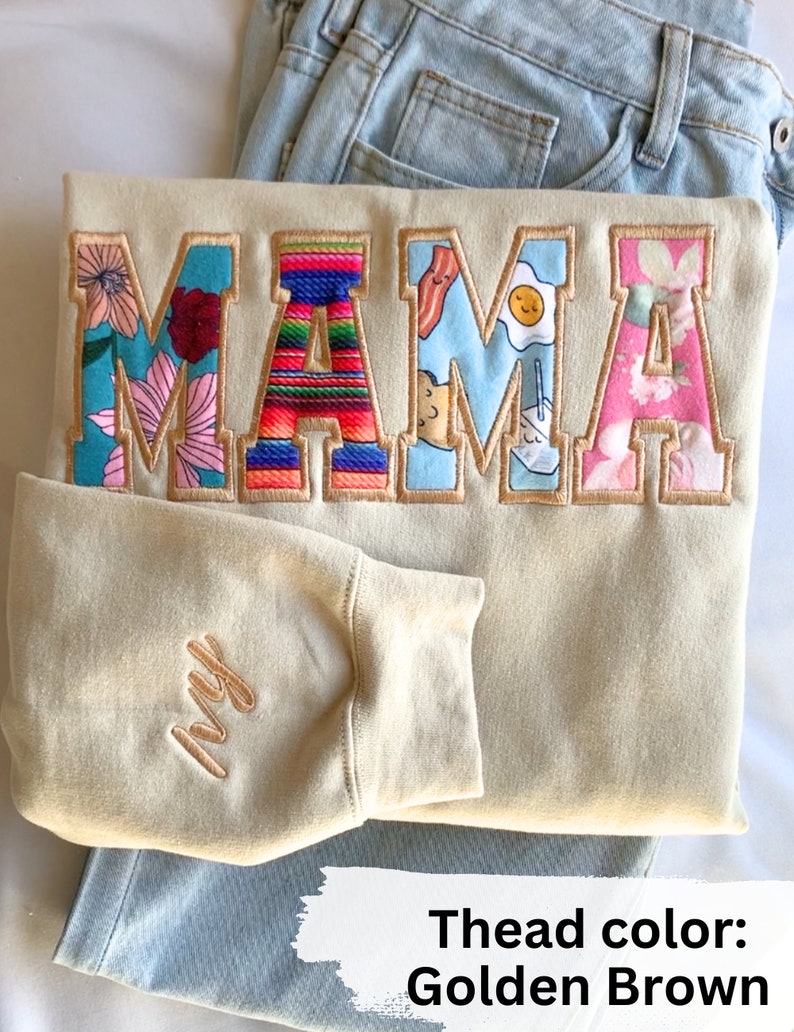 May include: A beige sweatshirt with the word "MAMA" embroidered in colorful patterned letters. The letters are outlined in golden brown thread. The sweatshirt has a small "Ivy" embroidered on the sleeve in golden brown thread.