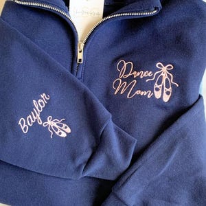 May include: Three sweatshirts with embroidered designs. The first sweatshirt is grey with purple embroidery that says "Dance Mom" with a dancer silhouette. The second sweatshirt is navy blue with pink embroidery that says "Dance Mom" with a pair of ballet shoes. The third sweatshirt is white with red embroidery that says "Brielle" with a pair of ballet shoes.