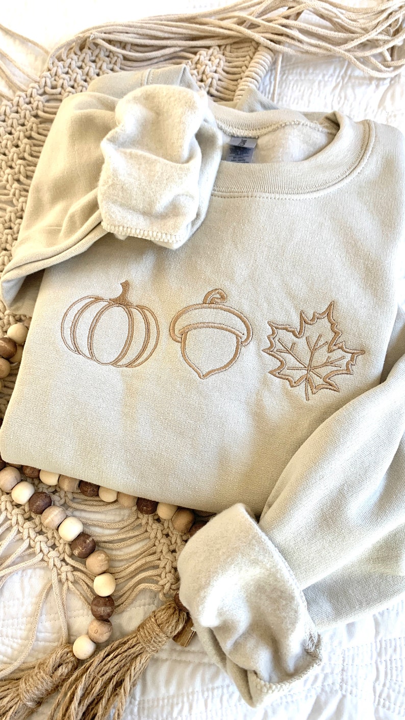 May include: A beige sweatshirt with embroidered images of a pumpkin, acorn, and maple leaf. The sweatshirt is folded and lying on a beige crocheted surface.