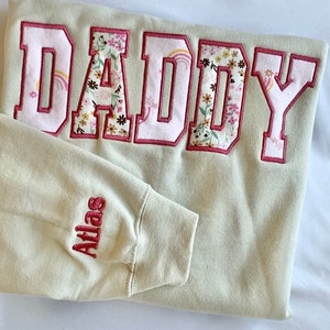 May include: A light beige sweatshirt with the word "DADDY" embroidered in large pink letters with a floral pattern inside. The word "Atlas" is embroidered in red on the left sleeve.