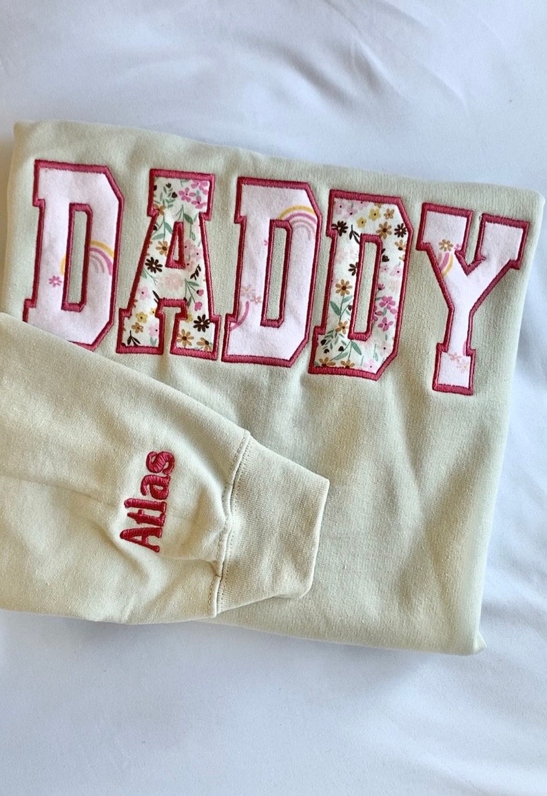 May include: A light beige sweatshirt with the word "DADDY" embroidered in large pink letters with a floral pattern inside. The word "Atlas" is embroidered in red on the left sleeve.
