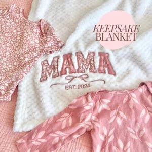Pode incluir: Uma manta de pelúcia branca com a palavra "MAMA" bordada em rosa com um padrão floral. A manta também tem o texto "EST. 2024" bordado abaixo da palavra "MAMA". A manta é cercada por duas roupas de bebê rosa com padrão floral.