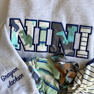 May include: A gray sweatshirt with a large, colorful "NINI" applique on the front. The applique is made of fabric with a dinosaur and other animal designs. The sweatshirt also has the name "Grayson Jackson" embroidered on the sleeve.
