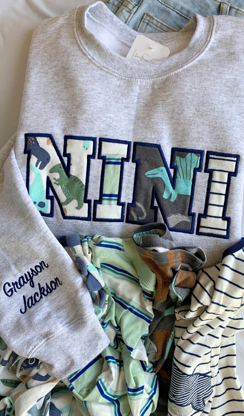May include: A gray sweatshirt with a large, colorful "NINI" applique on the front. The applique is made of fabric with a dinosaur and other animal designs. The sweatshirt also has the name "Grayson Jackson" embroidered on the sleeve.