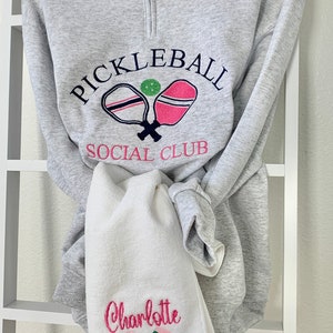 May include: Gray sweatshirt with a white and pink pickleball paddle design and the text "Pickleball Social Club". A white towel with the name "Charlotte" and a green and pink pickleball paddle design is hanging below the sweatshirt.