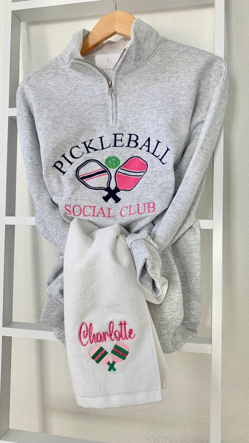 May include: Gray sweatshirt with a white and pink pickleball paddle design and the text "Pickleball Social Club". A white towel with the name "Charlotte" and a green and pink pickleball paddle design is hanging below the sweatshirt.