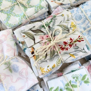 Puede incluir: Conjunto de paquetes de tela atados con hilo. La tela presenta varios patrones florales y bot&aacute;nicos en tonos azules, rosas, amarillos y verdes sobre un fondo blanco. Los paquetes est&aacute;n apilados.