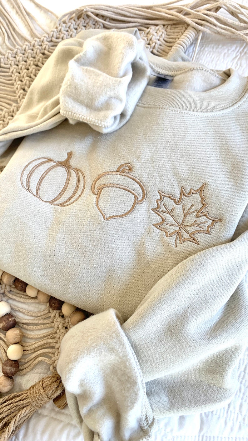 May include: A light beige sweatshirt with an embroidered design of a pumpkin, acorn, and maple leaf. The design is in a light brown color.