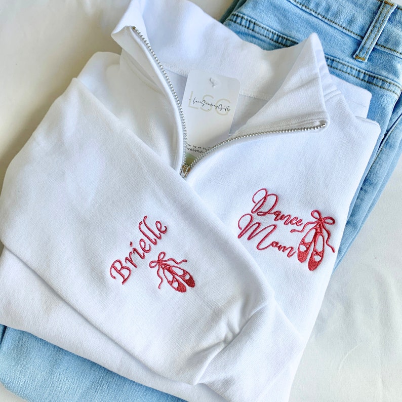May include: White zip-up sweatshirt with the text "Dance Mom" and a pair of pink ballet shoes embroidered on the front. The left sleeve has the name "Brielle" and a pair of pink ballet shoes embroidered on it.
