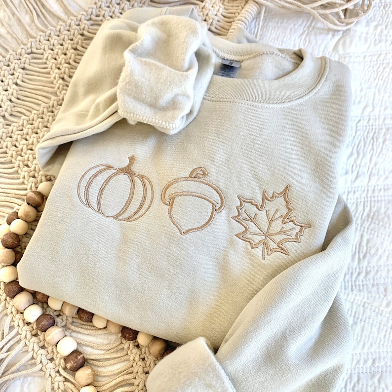May include: A beige sweatshirt with an embroidered pumpkin, acorn, and maple leaf design. The sweatshirt is folded and lying on a beige crocheted surface.