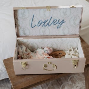 An open wooden trunk with a name label inside, containing various items such as a hairbrush and other personal belongings.