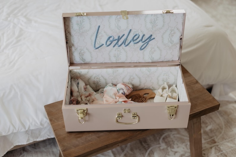 An open wooden trunk with a name label inside, containing various items such as a hairbrush and other personal belongings.