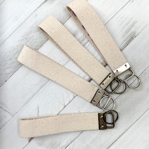 May include: Four canvas wrist keychains in a natural beige color. Each keychain has a metal clasp and a split ring for attaching keys. The keychains are arranged on a white wooden surface.