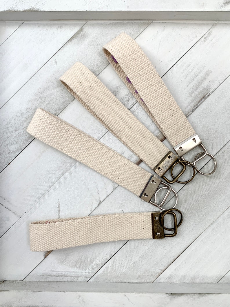 May include: Four canvas wrist keychains in a natural beige color. Each keychain has a metal clasp and a split ring for attaching keys. The keychains are arranged on a white wooden surface.