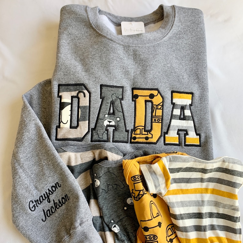 May include: Gray sweatshirt with the word "DADA" in large, colorful letters. The letters are decorated with images of animals and vehicles. The sweatshirt has a gray and white striped sleeve with the name "Grayson Jackson" embroidered in black.