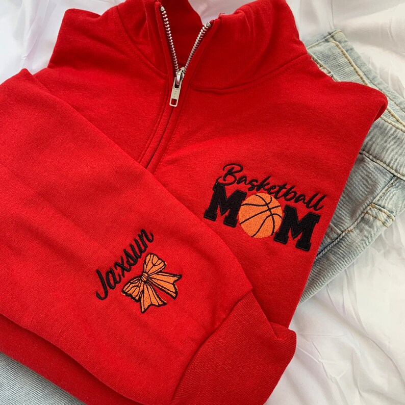 May include: Red zippered sweatshirt with the text "Basketball Mom" and a basketball graphic on the right side. The left side has the text "Jaxsun" and a bow graphic.