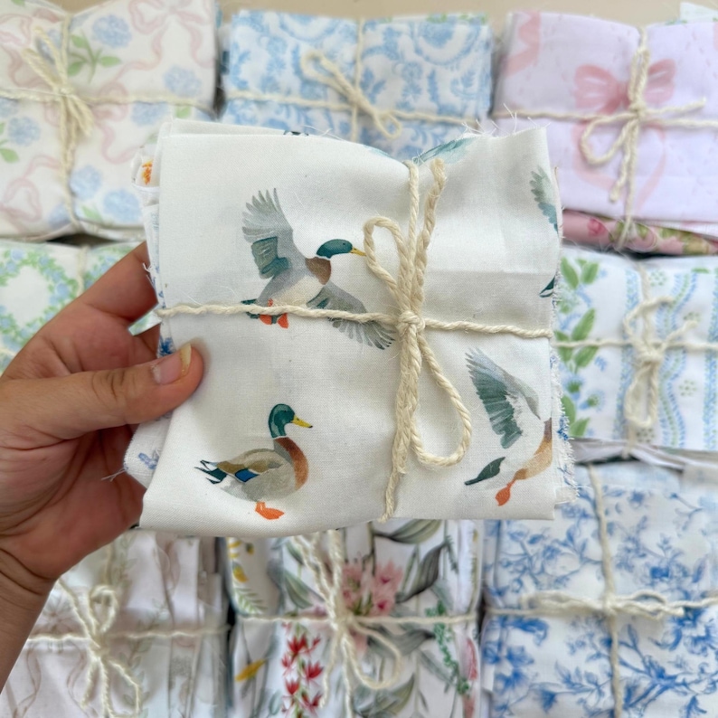 Puede incluir: Un paquete de cuadrados de tela atados con hilo. La tela superior presenta un dise&ntilde;o de patos en tonos azules, verdes y marrones sobre un fondo blanco. Otros paquetes muestran telas florales y estampadas en colores pastel.